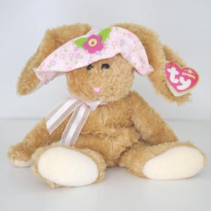 May include: A plush brown bunny with a pink floral hat and a striped ribbon bow. The bunny has cream-colored paws and a pink nose. A heart-shaped tag is attached to the ear.