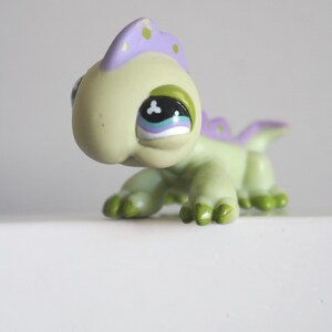 Puede incluir: Una pequeña figura de lagarto de color verde claro con una cabeza grande y ojos sobredimensionados. Presenta una cresta morada en la espalda con puntos verde claro, garras verde oscuro y un detalle ocular blanco con pupila negra.