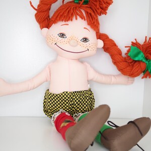 以下が含まれることがあります： 緑のリボンで結ばれた赤い糸の三つ編みを持つ手作りの人形。人形はそばかすのある笑顔の顔をしており、黄色と黒のチェック柄のショーツ、赤い靴下、茶色の靴を履いています。人形は座っています。