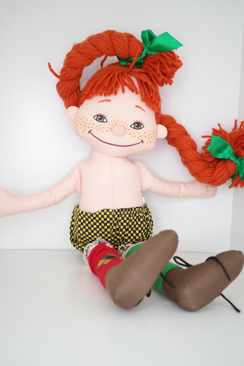 以下が含まれることがあります： 緑のリボンで結ばれた赤い糸の三つ編みを持つ手作りの人形。人形はそばかすのある笑顔の顔をしており、黄色と黒のチェック柄のショーツ、赤い靴下、茶色の靴を履いています。人形は座っています。