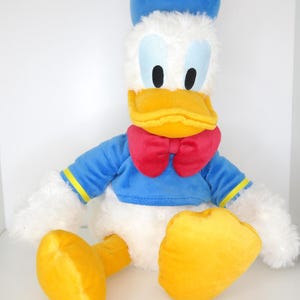 Puede incluir: Un peluche de Pato Donald, con una camisa de marinero azul, una pajarita roja y pies amarillos. El juguete tiene un cuerpo blanco, un pico amarillo y un sombrero azul. Los ojos son negros y el juguete está diseñado para parecerse al personaje de dibujos animados.