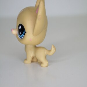 May include: A small, tan plastic toy dog with blue eyes and a pink nose. The dog is standing on a white surface.