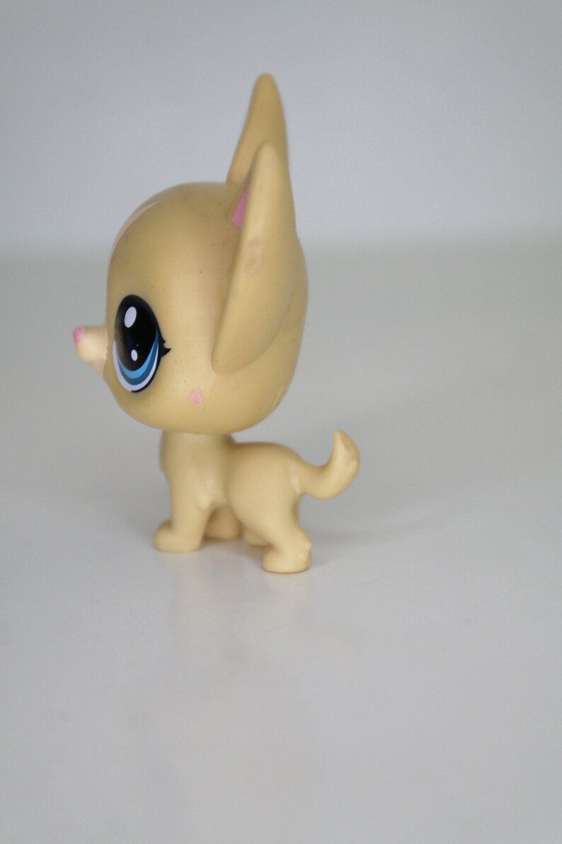 May include: A small, tan plastic toy dog with blue eyes and a pink nose. The dog is standing on a white surface.