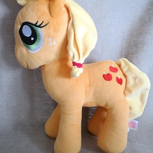 May include: A plush toy of an orange pony with a light yellow mane and tail. The pony features large green eyes, a red hair tie, and three embroidered red apple symbols on its flank. A small My Little Pony tag is attached to the rear leg.