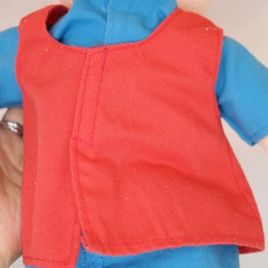K&ouml;nnte beinhalten: Puppe mit roter Weste &uuml;ber einem blauen Hemd. Die Puppe hat orangefarbene Z&ouml;pfe aus Garn. Die Weste hat ein schlichtes Design mit einer vertikalen Naht auf dem R&uuml;cken. Das blaue Hemd hat kurze &Auml;rmel. Die Haut der Puppe ist hellbeige.
