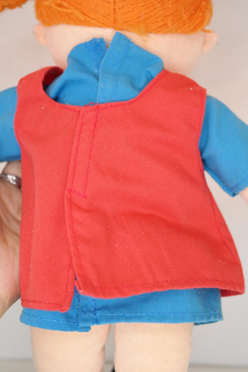 K&ouml;nnte beinhalten: Puppe mit roter Weste &uuml;ber einem blauen Hemd. Die Puppe hat orangefarbene Z&ouml;pfe aus Garn. Die Weste hat ein schlichtes Design mit einer vertikalen Naht auf dem R&uuml;cken. Das blaue Hemd hat kurze &Auml;rmel. Die Haut der Puppe ist hellbeige.