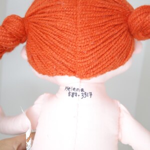 以下が含まれることがあります： オレンジ色の糸の三つ編みをした手作りの人形。人形はピンク色の体と黄色と黒のチェック柄のスカートをしています。「Helena」という名前と電話番号が人形の背中に書かれています。