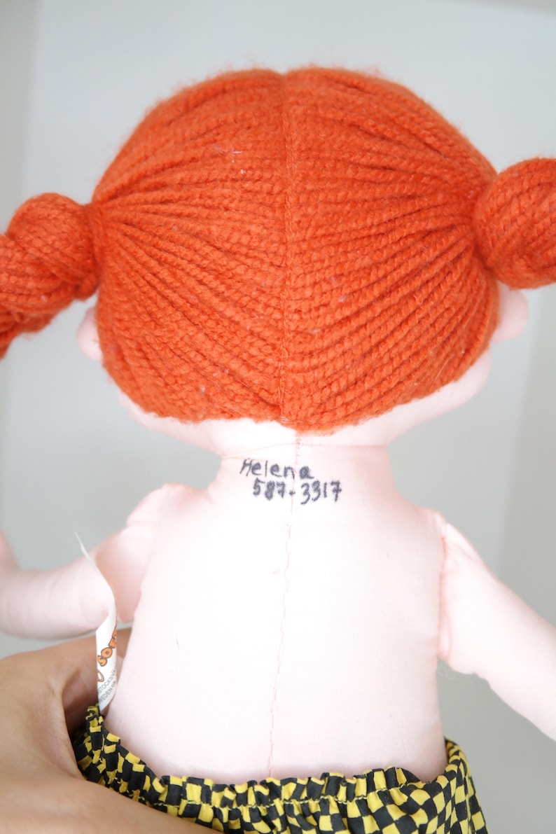 以下が含まれることがあります： オレンジ色の糸の三つ編みをした手作りの人形。人形はピンク色の体と黄色と黒のチェック柄のスカートをしています。「Helena」という名前と電話番号が人形の背中に書かれています。