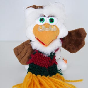 Puede incluir: Un juguete de peluche de búho con plumas blancas y marrones, ojos verdes y un pico naranja. Lleva una bufanda roja, verde y amarilla. El juguete tiene alas marrones y un chupete sujeto al pico.