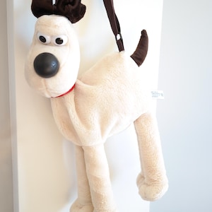 Puede incluir: Un bolso de hombro de peluche blanco con forma de perro con orejas marrones y un collar rojo. El bolso tiene una correa marrón y está hecho de un material suave y peludo.