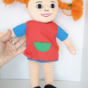 K&ouml;nnte beinhalten: Pl&uuml;schpuppe mit orangefarbenen Zopfz&ouml;pfen, einem roten Kleid mit gr&uuml;ner Tasche und schwarzen Schuhen. Die Puppe hat ein l&auml;chelndes Gesicht mit Sommersprossen und tr&auml;gt ein blaues Hemd unter dem Kleid. Die Puppe ist etwa 30 cm gro&szlig;.