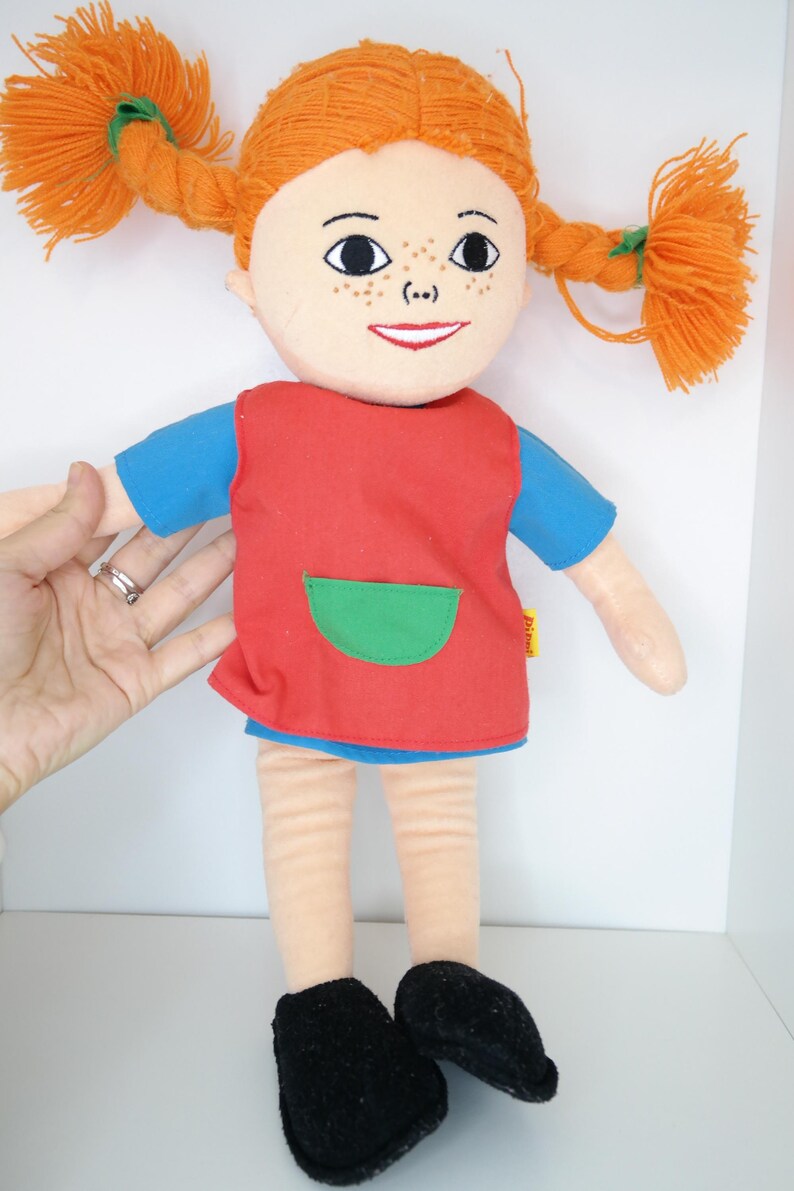 K&ouml;nnte beinhalten: Pl&uuml;schpuppe mit orangefarbenen Zopfz&ouml;pfen, einem roten Kleid mit gr&uuml;ner Tasche und schwarzen Schuhen. Die Puppe hat ein l&auml;chelndes Gesicht mit Sommersprossen und tr&auml;gt ein blaues Hemd unter dem Kleid. Die Puppe ist etwa 30 cm gro&szlig;.