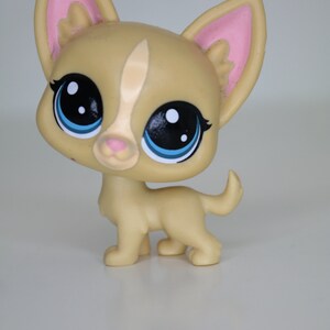 May include: A small, tan-colored plastic toy dog with large blue eyes and pink ears. The dog is standing on a white surface.