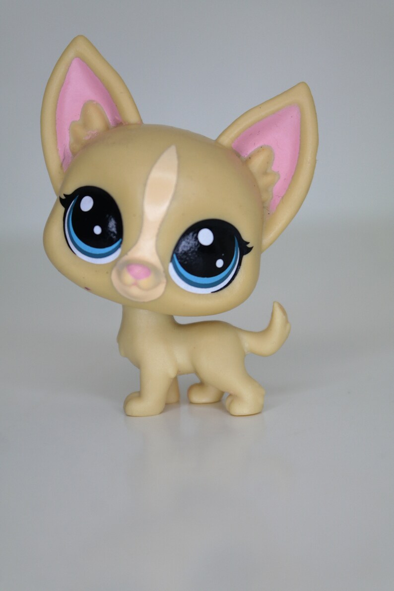 May include: A small, tan-colored plastic toy dog with large blue eyes and pink ears. The dog is standing on a white surface.