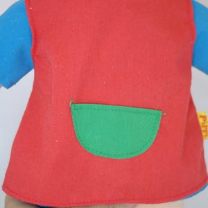 K&ouml;nnte beinhalten: Nahaufnahme einer Puppe, die eine rote Weste mit einer gr&uuml;nen Tasche &uuml;ber einem blauen Langarmshirt tr&auml;gt. Die Puppe hat einen hellen Hautton und orangefarbene Haare. Die Weste hat eine abgerundete gr&uuml;ne Tasche.