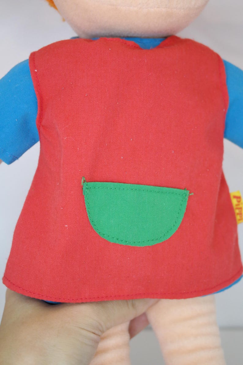 K&ouml;nnte beinhalten: Nahaufnahme einer Puppe, die eine rote Weste mit einer gr&uuml;nen Tasche &uuml;ber einem blauen Langarmshirt tr&auml;gt. Die Puppe hat einen hellen Hautton und orangefarbene Haare. Die Weste hat eine abgerundete gr&uuml;ne Tasche.