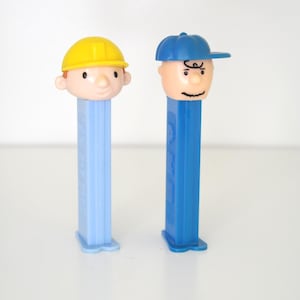 Puede incluir: Dos dispensadores de PEZ. Uno tiene un casco amarillo y el otro una gorra de béisbol azul. Ambos tienen cuerpos de color azul claro y están de pie sobre una superficie blanca. Los dispensadores son artículos novedosos.
