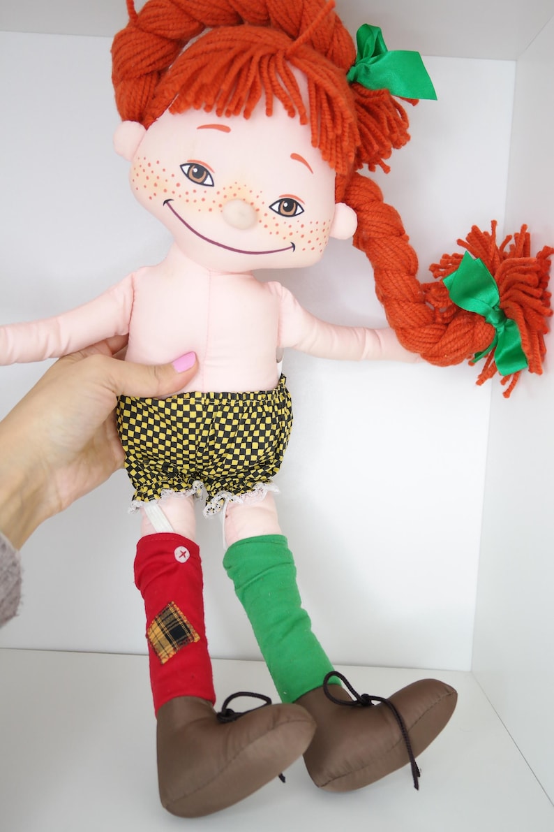 以下が含まれることがあります： 赤い糸の髪を三つ編みにし、緑のリボンをつけた手作りの人形。そばかすのある顔で、黄と黒のチェック柄のショートパンツ、赤と緑の靴下、茶色の靴を履いています。ユニークで気まぐれな玩具。