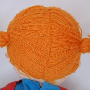 K&ouml;nnte beinhalten: Eine Puppe mit leuchtend orangefarbenen Z&ouml;pfen aus Garn. Die Puppe tr&auml;gt ein blaues Hemd und eine rote Weste. Das Haar der Puppe ist zu zwei Z&ouml;pfen gestylt, einer auf jeder Seite des Kopfes. Das Gesicht der Puppe ist nicht sichtbar.