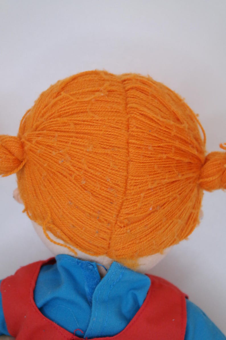 K&ouml;nnte beinhalten: Eine Puppe mit leuchtend orangefarbenen Z&ouml;pfen aus Garn. Die Puppe tr&auml;gt ein blaues Hemd und eine rote Weste. Das Haar der Puppe ist zu zwei Z&ouml;pfen gestylt, einer auf jeder Seite des Kopfes. Das Gesicht der Puppe ist nicht sichtbar.
