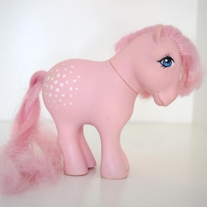 Könnte beinhalten: Ein rosa Spielzeugpony mit fließender rosa Mähne und Schweif. Das Pony hat blaue Augen und weiße Tupfen an der Seite. Das Spielzeug steht auf einer weißen Oberfläche.