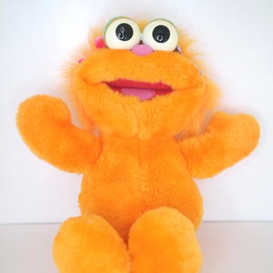 Puede incluir: Juguete de peluche naranja con ojos grandes, una nariz rosa y una lengua rosa que sobresale. El juguete tiene un lazo rosa en la cabeza y está sentado sobre una superficie blanca.