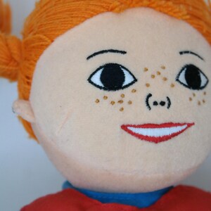 K&ouml;nnte beinhalten: Eine Pl&uuml;schpuppe mit orangefarbenem Garnhaar, das zu Z&ouml;pfen geflochten ist. Die Puppe hat einen hellen Hautton, gestickte schwarze Augen, Sommersprossen und einen roten, l&auml;chelnden Mund. Sie tr&auml;gt ein rotes Oberteil &uuml;ber einem blauen Hemd.
