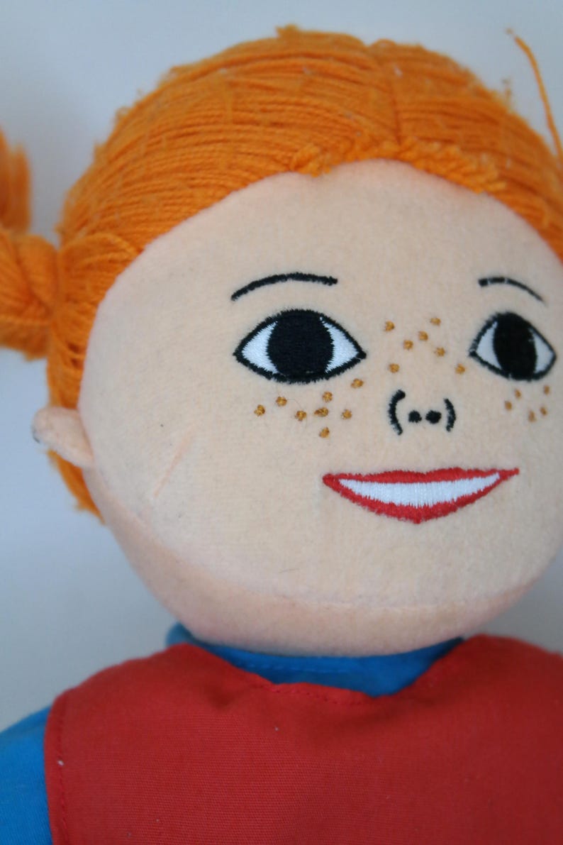 K&ouml;nnte beinhalten: Eine Pl&uuml;schpuppe mit orangefarbenem Garnhaar, das zu Z&ouml;pfen geflochten ist. Die Puppe hat einen hellen Hautton, gestickte schwarze Augen, Sommersprossen und einen roten, l&auml;chelnden Mund. Sie tr&auml;gt ein rotes Oberteil &uuml;ber einem blauen Hemd.
