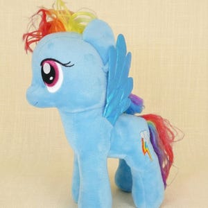 May include: A light blue plush pony toy featuring a rainbow-colored mane and tail, large pink eyes, and metallic blue wings. A small cloud and lightning bolt emblem is embroidered on the flank of the soft, stuffed four-legged creature.