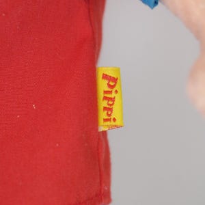 K&ouml;nnte beinhalten: Nahaufnahme eines roten Hemdes mit blauen &Auml;rmeln. Ein gelbes Etikett mit dem Wort "Pippi" in wei&szlig;er Schrift ist am Hemd befestigt. Das Hemd ist aus einem weichen Material.