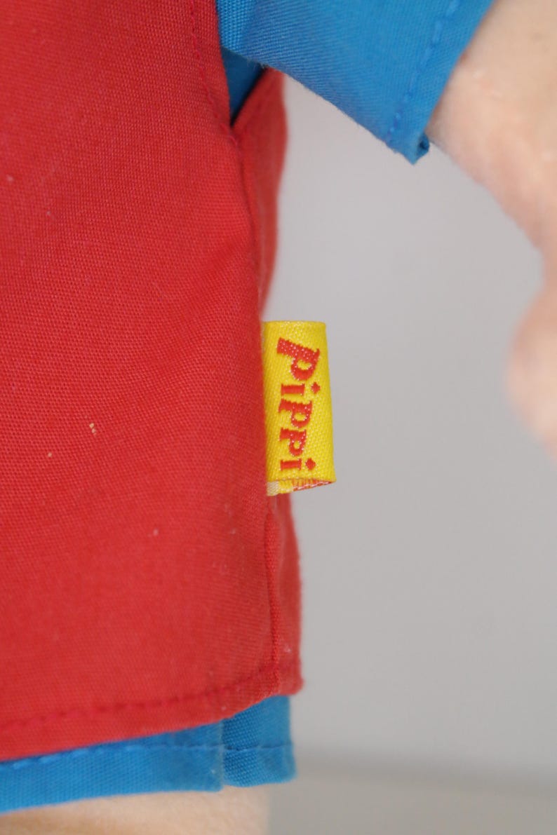 K&ouml;nnte beinhalten: Nahaufnahme eines roten Hemdes mit blauen &Auml;rmeln. Ein gelbes Etikett mit dem Wort "Pippi" in wei&szlig;er Schrift ist am Hemd befestigt. Das Hemd ist aus einem weichen Material.