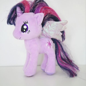 Puede incluir: Un peluche de pony lavanda con una larga melena y cola que fluyen en tonos de morado, rosa y azul. El pony tiene alas iridiscentes, un cuerno y una marca de belleza en forma de estrella. Es un juguete para niños.