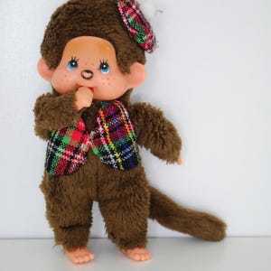 Peut inclure: Une poupée singe en peluche marron avec un visage couleur pêche, des yeux bleus et un pouce dans la bouche. La poupée porte un gilet à carreaux rouges, verts et noirs ainsi qu'un béret assorti. Elle a une longue queue et une texture douce et duveteuse.