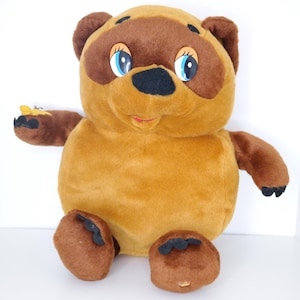 Peut inclure: Une peluche en forme d'ours avec une fourrure marron, des accents marron foncé autour des yeux et des oreilles, et un nez noir. L'ours a des yeux bleus et une petite bouche rouge. Il tient un petit objet jaune et noir dans sa patte gauche.