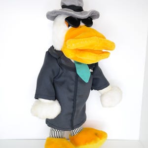 Könnte beinhalten: Ein Plüschtier, das eine Ente darstellt, die einen grauen Fedora-Hut, eine schwarze Sonnenbrille und einen dunkelgrauen Anzug mit einer türkisfarbenen Krawatte trägt. Die Ente hat einen großen orangen Schnabel und gelbe Füße mit einer schwarz-weiß gestreiften Hose.