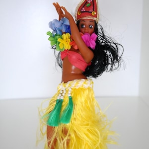 Puede incluir: Muñeca vestida con un atuendo tradicional hawaiano de hula. La muñeca tiene piel oscura, pelo largo y negro, y está adornada con un tocado rojo, leis coloridos y una falda amarilla con borlas verdes. La muñeca está en posición de baile.