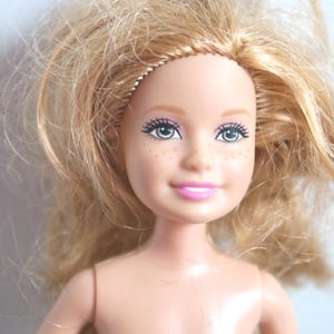 May include: A close-up of a doll's face with blonde hair and green eyes. The doll has painted eyelashes, eyebrows, and pink lips. Small freckles are visible on the doll's face. The doll's skin is a light peach color.