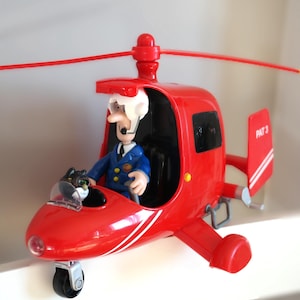 Pode incluir: Um helicóptero de brinquedo vermelho brilhante com uma figura de uniforme azul e um gato preto. O helicóptero tem uma faixa branca e o texto "PAT 3" na cauda. Possui uma única roda dianteira e uma hélice vermelha, um brinquedo infantil.