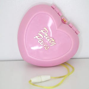 Può includere: Un giocattolo rosa a forma di cuore con la scritta dorata "Pretty Pixies". Il giocattolo ha un cordino giallo e una chiusura bianca. Il giocattolo è chiuso e ha una finitura liscia e lucida.