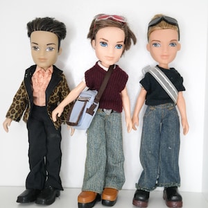 May include: Three fashion dolls standing side by side. The left doll wears a leopard print jacket, peach ruffled shirt, and black trousers. The center doll has a maroon sweater vest, blue jeans, and a light blue crossbody bag. The right doll wears a black t-shirt, blue jeans, and a silver crossbody bag.