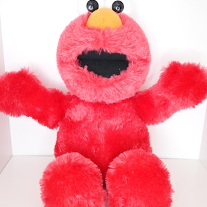 Puede incluir: Un peluche rojo con grandes ojos blancos, una nariz amarilla y una boca negra ancha. El juguete tiene los brazos extendidos y pies grandes y esponjosos. El fondo es blanco.