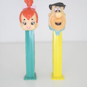 Puede incluir: Dos figuras de juguete de plástico vintage de personajes de la caricatura Los Picapiedra. Las figuras están unidas a la parte superior de dispensadores de plástico amarillo y turquesa. Las figuras son de Fred Picapiedra y Wilma Picapiedra.