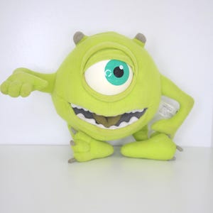 Puede incluir: Un peluche del personaje Mike Wazowski de la película de animación Monstruos, S.A. El monstruo verde lima tiene un ojo grande con iris blanco y pupila turquesa. Tiene pequeños cuernos y una amplia sonrisa sin dientes.