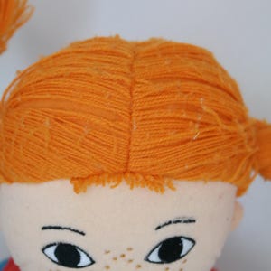 K&ouml;nnte beinhalten: Nahaufnahme eines Puppengesichts mit orangefarbenem Garnhaar, das zu Z&ouml;pfen gestylt ist. Die Puppe hat einen hellen Teint, schwarze Augen, Sommersprossen und ein rotes Hemd. Der Ausdruck der Puppe ist ein leichtes L&auml;cheln.