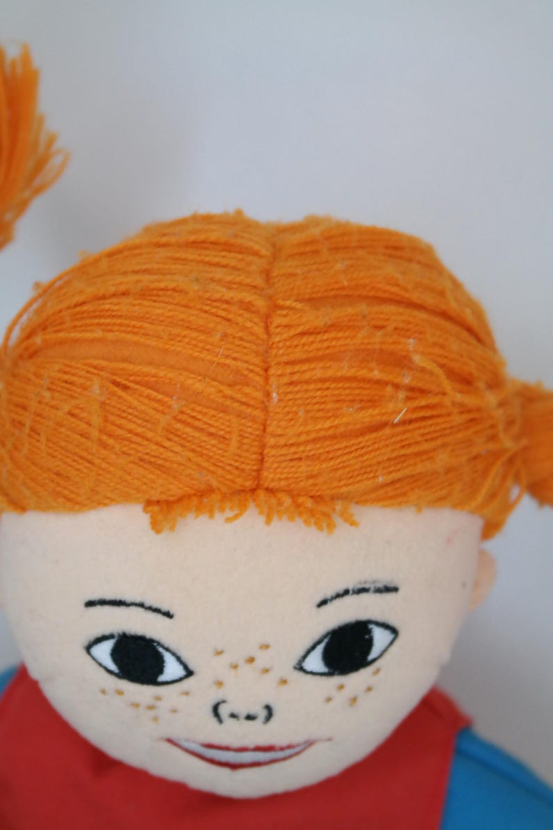 K&ouml;nnte beinhalten: Nahaufnahme eines Puppengesichts mit orangefarbenem Garnhaar, das zu Z&ouml;pfen gestylt ist. Die Puppe hat einen hellen Teint, schwarze Augen, Sommersprossen und ein rotes Hemd. Der Ausdruck der Puppe ist ein leichtes L&auml;cheln.