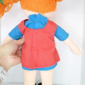 K&ouml;nnte beinhalten: Handgemachte Puppe mit orangefarbenen Z&ouml;pfen aus Garn, die ein blaues Hemd und eine rote Weste tr&auml;gt. Die Puppe hat schwarze Schuhe und wird von einer Hand gehalten. Das Haar der Puppe ist zu zwei Z&ouml;pfen geflochten.