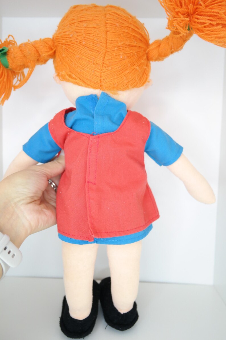 K&ouml;nnte beinhalten: Handgemachte Puppe mit orangefarbenen Z&ouml;pfen aus Garn, die ein blaues Hemd und eine rote Weste tr&auml;gt. Die Puppe hat schwarze Schuhe und wird von einer Hand gehalten. Das Haar der Puppe ist zu zwei Z&ouml;pfen geflochten.