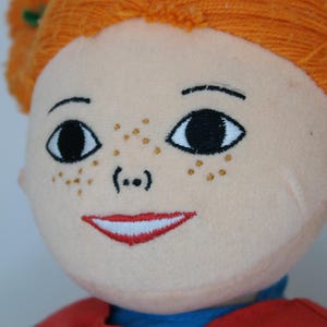 K&ouml;nnte beinhalten: Stoffpuppe mit orangefarbenem Garnhaar, das zu Z&ouml;pfen geflochten ist. Die Puppe hat gestickte Gesichtsz&uuml;ge, darunter schwarze Augen, Sommersprossen und einen roten, l&auml;chelnden Mund. Sie tr&auml;gt ein rotes Oberteil &uuml;ber einem blauen Hemd.