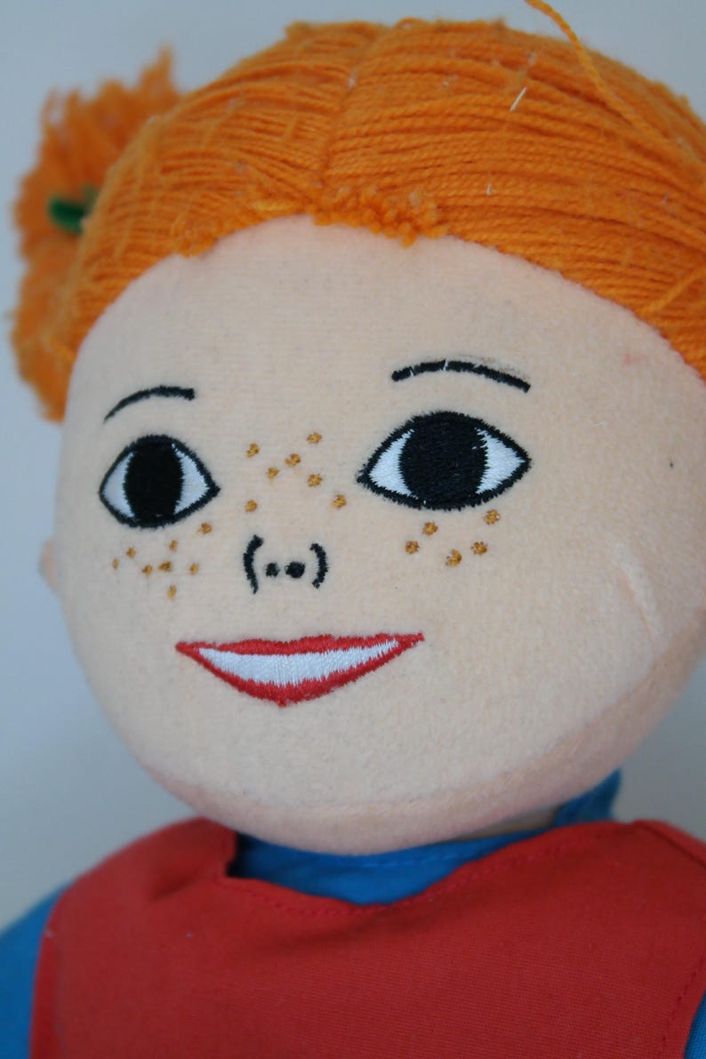 K&ouml;nnte beinhalten: Stoffpuppe mit orangefarbenem Garnhaar, das zu Z&ouml;pfen geflochten ist. Die Puppe hat gestickte Gesichtsz&uuml;ge, darunter schwarze Augen, Sommersprossen und einen roten, l&auml;chelnden Mund. Sie tr&auml;gt ein rotes Oberteil &uuml;ber einem blauen Hemd.