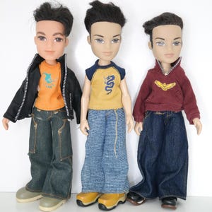 Könnte beinhalten: Drei Modepuppen mit dunklem Haar und stilvollen Outfits. Die Puppe links trägt eine schwarze Jacke, ein orangefarbenes Hemd mit Grafik und Jeans. Die mittlere Puppe hat ein gelbes Hemd, blaue Jeans und gelbe Schuhe. Die rechte Puppe trägt einen weinroten Kapuzenpullover und dunkelblaue Jeans.